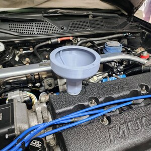 May include: A grey plastic funnel with a wide opening and a narrow spout, sitting on top of a black engine block with blue spark plug wires. The engine block has the word "MUGEN" printed on it.