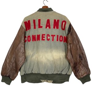 Puede incluir: Chaqueta bomber vintage con mangas de cuero marrón y cuerpo gris descolorido. La parte trasera presenta las palabras "MILANO CONNECTION" en letras rojas. La chaqueta tiene cuello, puños y cintura verdes, colgada de una percha de madera.