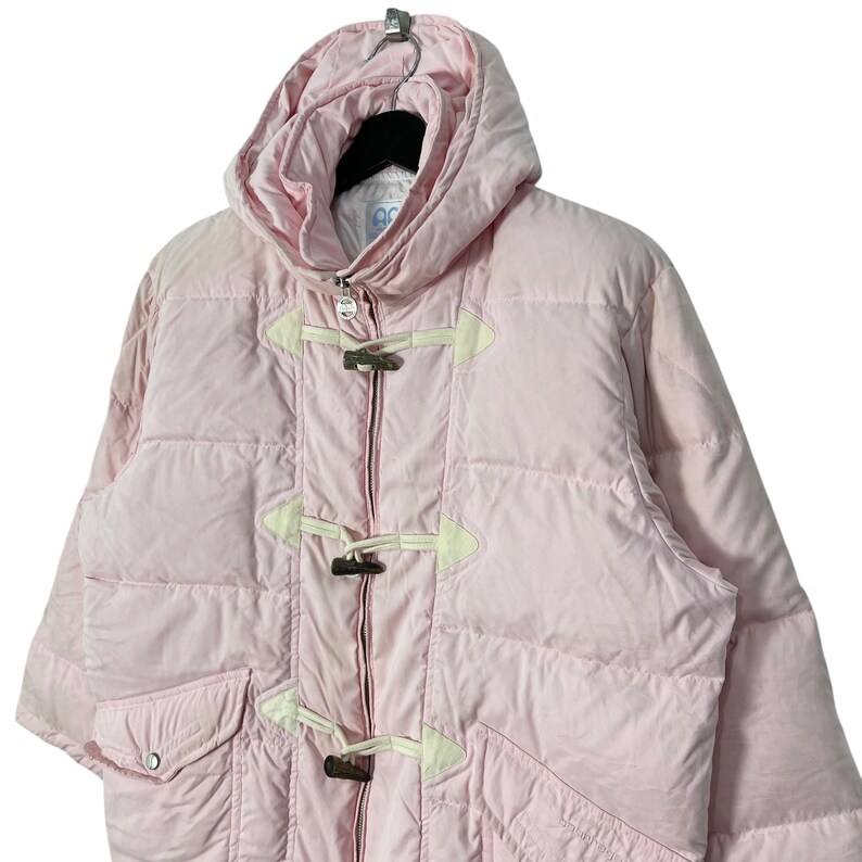 K&ouml;nnte beinhalten: Eine rosafarbene Steppjacke mit Kapuze und Knebelverschl&uuml;ssen. Die Jacke hat ein gestepptes Design und einen Rei&szlig;verschluss.