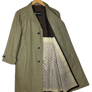 Puede incluir: Una gabardina larga de color verde oliva con cierre de botones negros y forro estampado. El forro presenta un diseño geométrico y la palabra "LANVIN" en letras negras.