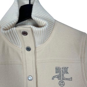 May include: Cream-colored jacket with a ribbed knit collar and cuffs. The jacket features a button-up front and a rhinestone emblem on the chest. The jacket is made of a soft, textured material.