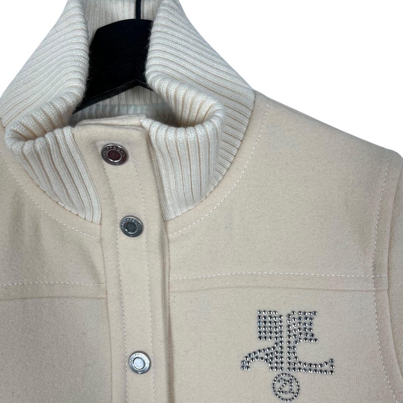 May include: Cream-colored jacket with a ribbed knit collar and cuffs. The jacket features a button-up front and a rhinestone emblem on the chest. The jacket is made of a soft, textured material.