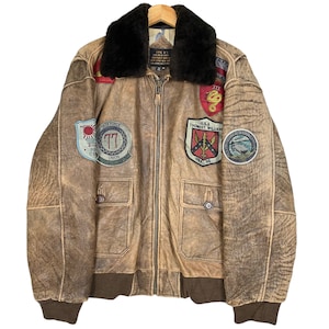 May include: A brown leather bomber jacket with a faux fur collar and multiple patches. The patches include a red, white and blue patch with the text "U.S.A. THOMAS WILLIAMS" and "HMX-36", a patch with a red, white and blue snake, a patch with a red circle and the text "TASK FORCE 77", and a patch with a blue circle and the text "STRIKE FIGHTER CENTER UNION".