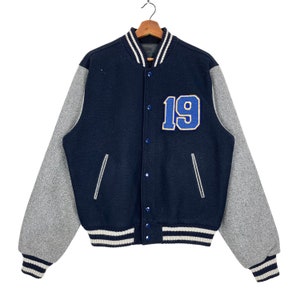Puede incluir: Una chaqueta de béisbol azul marino y gris con rayas blancas y azul marino en los puños y la cintura. La chaqueta tiene un gran "19" azul y amarillo en la parte delantera.