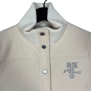 May include: Cream-colored jacket with a ribbed, off-white collar and three silver snap closures. The jacket features a stitched design and a rhinestone emblem on the front. The jacket is hanging on a black hanger.