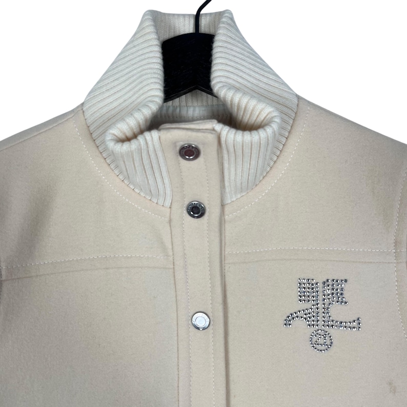 May include: Cream-colored jacket with a ribbed, off-white collar and three silver snap closures. The jacket features a stitched design and a rhinestone emblem on the front. The jacket is hanging on a black hanger.