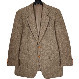 Peut inclure: Un blazer en tweed marron avec une fermeture à deux boutons et des revers crantés. Le blazer a deux poches avant et trois boutons sur chaque manchette. La doublure intérieure est de couleur marron clair. Le blazer est suspendu à un cintre noir.