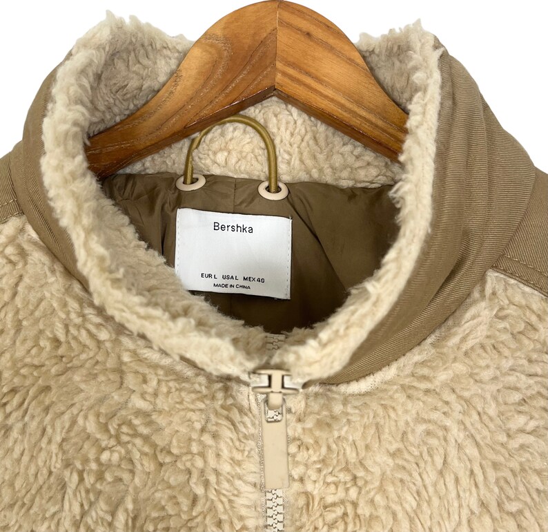 May include: A beige sherpa jacket with a brown collar and zipper closure. The jacket has a Bershka label with the text "EUR L USAL MEX 40 MADE IN CHINA".