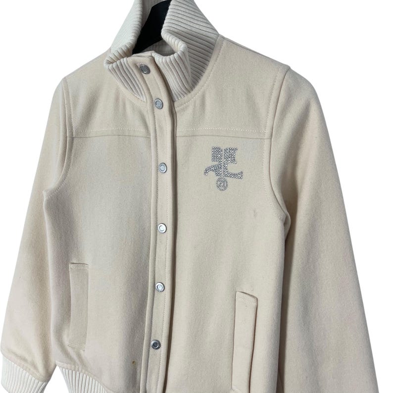 May include: Cream-colored varsity jacket with a ribbed collar and cuffs. The jacket features a button-up front, two front pockets, and a rhinestone emblem on the chest. The jacket has a classic, sporty design.