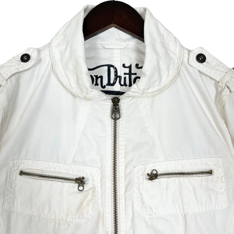 K&ouml;nnte beinhalten: Wei&szlig;e Baumwolljacke mit Rei&szlig;verschluss und mehreren Taschen. Die Jacke hat ein "Von Dutch" Logo auf der Innenseite des Kragens.
