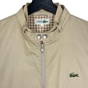 May include: A beige zip-up jacket with a brown and white checkered lining. The jacket has a small green crocodile logo on the left chest and the text "CHEMISELACOSTEL" on the lining.