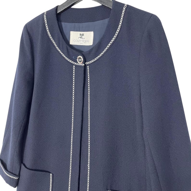 May include: A navy blue blazer with a white chain trim around the neckline and down the front. The blazer has a single button closure and a small pocket on the left side. The label "JC Courr&egrave;ges" is visible on the inside of the blazer.