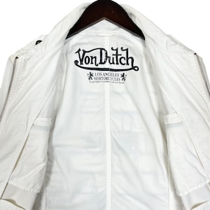 K&ouml;nnte beinhalten: Wei&szlig;e Baumwolljacke mit Rei&szlig;verschluss. Die Jacke hat ein schwarzes gesticktes Logo mit der Aufschrift "Von Dutch Los Angeles Motorcycles Everything's Going To Be All Right."