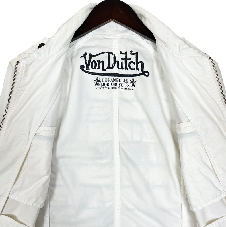 K&ouml;nnte beinhalten: Wei&szlig;e Baumwolljacke mit Rei&szlig;verschluss. Die Jacke hat ein schwarzes gesticktes Logo mit der Aufschrift "Von Dutch Los Angeles Motorcycles Everything's Going To Be All Right."
