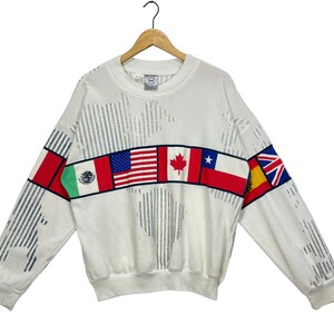 Può includere: Felpa bianca con un design a mappa del mondo e una fascia di bandiere sul petto. Le bandiere includono Messico, Stati Uniti, Canada, Cile, Spagna e Regno Unito.