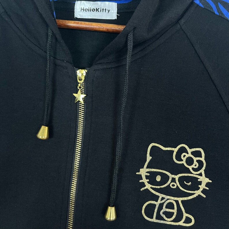May include: Black zip-up hoodie with a gold Hello Kitty graphic and a gold star zipper pull. The hoodie has blue and black zebra stripes on the sleeves.