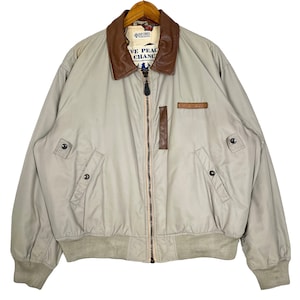 May include: A beige bomber jacket with a brown leather collar and a zipper closure. The jacket has two pockets on the front and a patch on the left chest that says "Give Peace a Chance".
