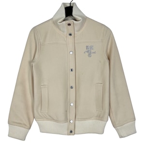 May include: Cream-colored varsity jacket with a ribbed collar, cuffs, and hem. The jacket features a button-up front, two front pockets, and an embroidered design on the chest. The jacket's design includes the text "Best Friend" in a stylized font.