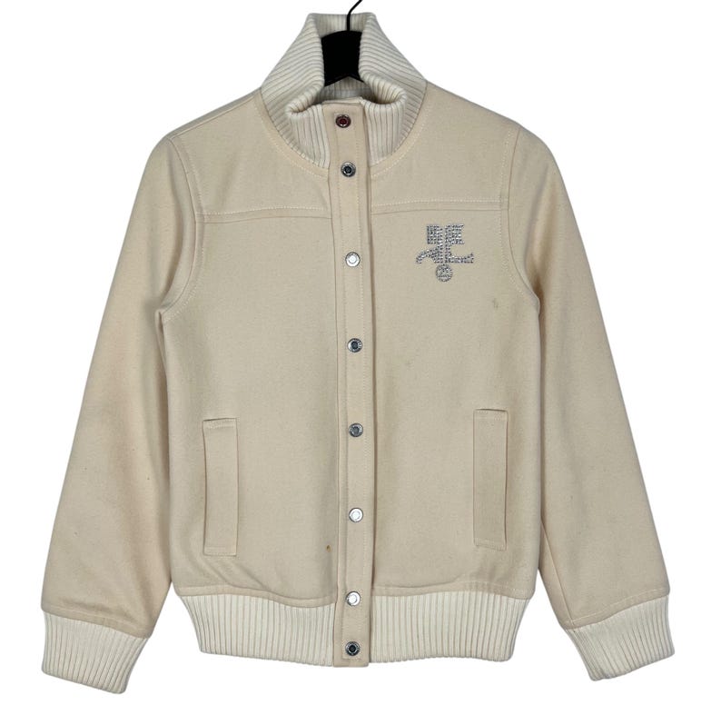 May include: Cream-colored varsity jacket with a ribbed collar, cuffs, and hem. The jacket features a button-up front, two front pockets, and an embroidered design on the chest. The jacket's design includes the text "Best Friend" in a stylized font.