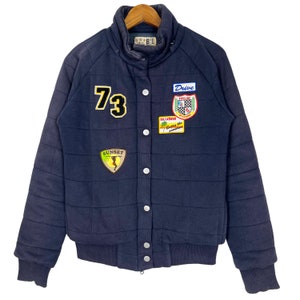 Puede incluir: Chaqueta acolchada azul marino con múltiples parches. La chaqueta presenta un parche "73", un parche "Drive" y un parche "Sunset". Tiene cuello y puños de canalé, y cierre de botones.