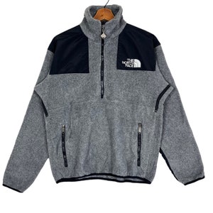 May include: A gray fleece pullover with a black panel on the chest and a zipper up the front. The zipper has a pull tab with a small logo. The logo on the chest says "THE NORTH FACE".