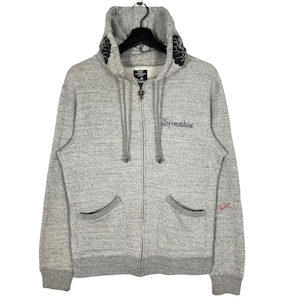 Op de afbeelding: Een lichtgrijze hoodie met ritssluiting, capuchon met trekkoord en twee voorzakken. De hoodie heeft de geborduurde tekst "Sofemachine" op de borst en bloemmotieven op de capuchon. De mouwen en zoom zijn donkerder grijs.