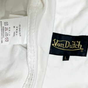 K&ouml;nnte beinhalten: Wei&szlig;es Baumwollhemd mit einem schwarzen Etikett, auf dem "Von Dutch" in goldenen Buchstaben steht. Das Etikett tr&auml;gt auch die Gr&ouml;&szlig;e "L". Das Hemd hat ein Pflegeetikett mit Anweisungen in Japanisch und Englisch.