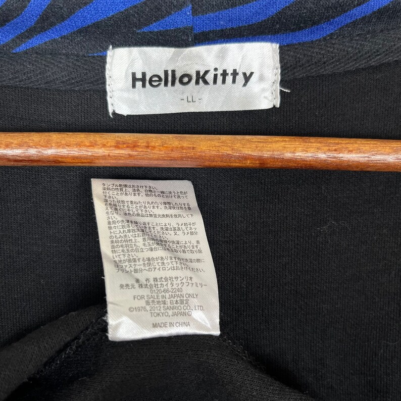 May include: Black Hello Kitty tag with white text that reads "Hello Kitty -LL-" and care instructions in Japanese. The tag is attached to a black garment.