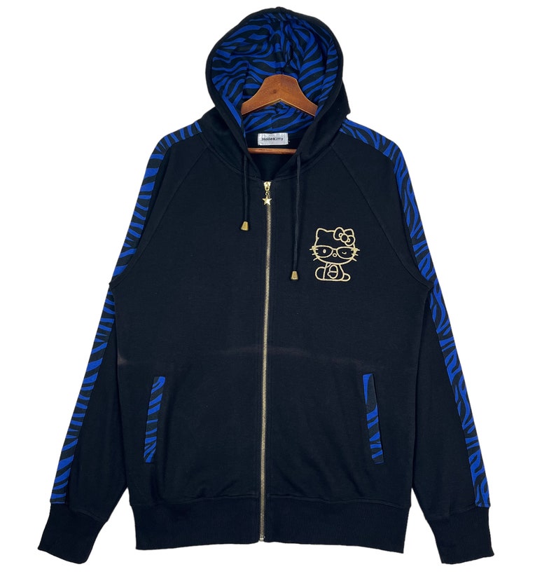 May include: Black zip-up hoodie with blue and black zebra print on the sleeves and hood. The hoodie has a gold Hello Kitty graphic on the front.