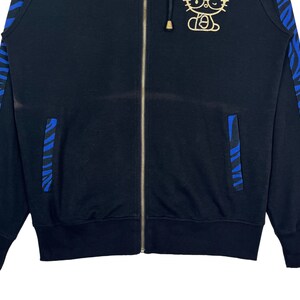 May include: Black zippered hoodie with blue and white striped trim on the sleeves and pockets. The hoodie features a gold Hello Kitty graphic on the front.