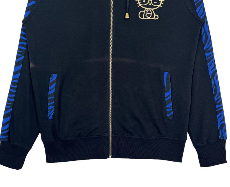 May include: Black zippered hoodie with blue and white striped trim on the sleeves and pockets. The hoodie features a gold Hello Kitty graphic on the front.