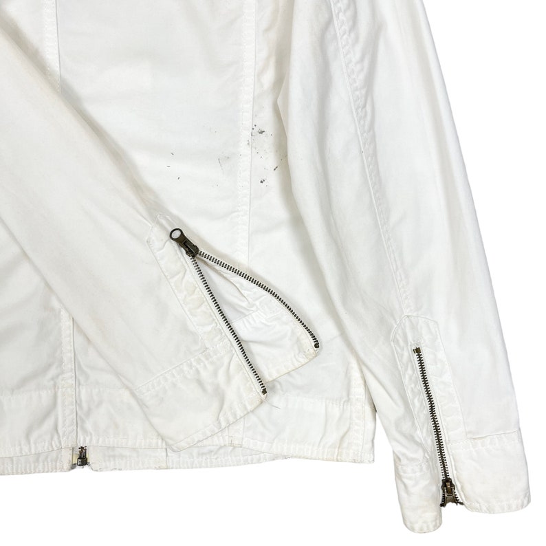 K&ouml;nnte beinhalten: Wei&szlig;e Baumwolljacke mit Rei&szlig;verschluss-&Auml;rmelb&uuml;ndchen und Rei&szlig;verschluss. Die Jacke hat einen leicht abgenutzten Look.