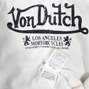 K&ouml;nnte beinhalten: Wei&szlig;es Von Dutch Los Angeles Motorcycles T-Shirt mit schwarzer Schrift und einem L&ouml;wenlogo. Der Text "Everything's going to be all right." ist unter dem Logo gedruckt.