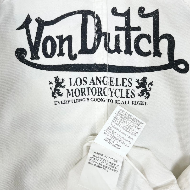 K&ouml;nnte beinhalten: Wei&szlig;es Von Dutch Los Angeles Motorcycles T-Shirt mit schwarzer Schrift und einem L&ouml;wenlogo. Der Text "Everything's going to be all right." ist unter dem Logo gedruckt.