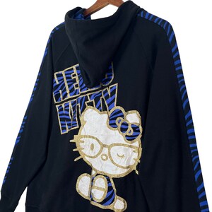 May include: Black hoodie with blue and white striped trim. The hoodie features a Hello Kitty graphic with gold accents and the text "Hello Kitty" in gold lettering.