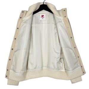 May include: Cream-colored varsity jacket with a ribbed collar, cuffs, and hem. The jacket features a button-up front and a white satin lining. The inside label shows the brand logo. A classic and stylish outerwear piece.