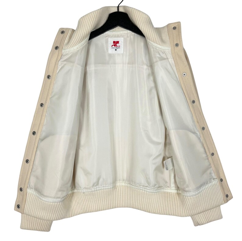 May include: Cream-colored varsity jacket with a ribbed collar, cuffs, and hem. The jacket features a button-up front and a white satin lining. The inside label shows the brand logo. A classic and stylish outerwear piece.