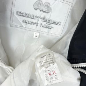 Może przedstawiać: Zbliżenie na wnętrze białej kurtki z czarnym wykończeniem. Kurtka ma markę "courr&egrave;ges" i napis "sport futur". Mniejsza metka pokazuje rozmiar "9". Inna metka pokazuje skład materiału i instrukcje pielęgnacji.