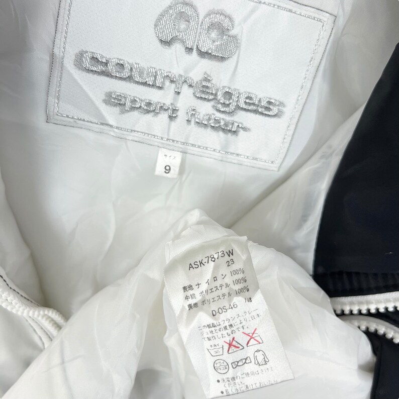 Może przedstawiać: Zbliżenie na wnętrze białej kurtki z czarnym wykończeniem. Kurtka ma markę "courr&egrave;ges" i napis "sport futur". Mniejsza metka pokazuje rozmiar "9". Inna metka pokazuje skład materiału i instrukcje pielęgnacji.