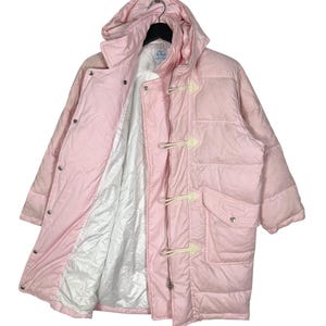 K&ouml;nnte beinhalten: Eine rosafarbene Steppjacke mit Kapuze und einem Knebelverschluss. Die Jacke hat ein wei&szlig;es Futter und einen Aufn&auml;her mit dem Text "CC" auf der Innenseite.