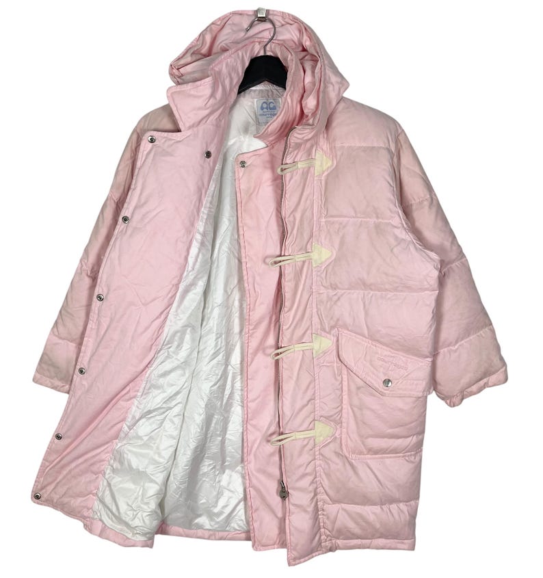 K&ouml;nnte beinhalten: Eine rosafarbene Steppjacke mit Kapuze und einem Knebelverschluss. Die Jacke hat ein wei&szlig;es Futter und einen Aufn&auml;her mit dem Text "CC" auf der Innenseite.