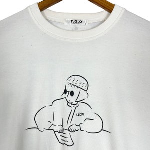 以下が含まれることがあります： 帽子とサングラスをかけた人の黒い線画が描かれた白いTシャツ。その人はテーブルに座って紙に書いています。Tシャツには「LEON」という文字が書かれています。