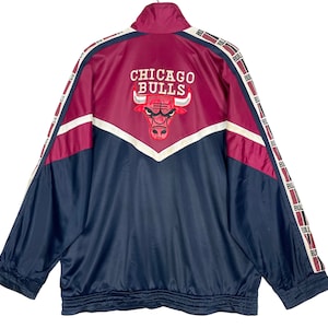 May include: A navy blue and burgundy windbreaker jacket with white piping and the Chicago Bulls logo on the back. The jacket has a zipper closure and long sleeves.