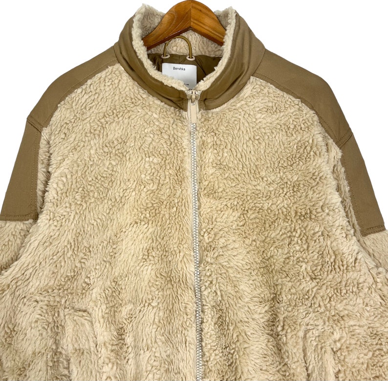 May include: A beige sherpa jacket with brown corduroy accents on the shoulders and sleeves. The jacket has a zipper closure and a stand-up collar.