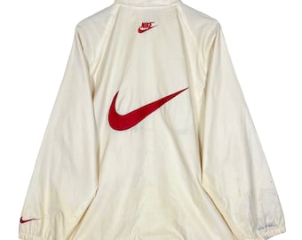 Pode incluir: Uma jaqueta corta-vento Nike branca com um logotipo Nike swoosh vermelho nas costas. A jaqueta tem um fecho de correr e uma gola alta.