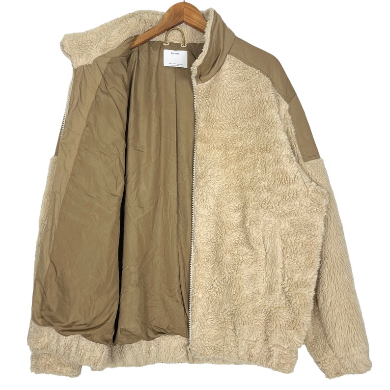 May include: A beige and brown fleece jacket with a zipper closure. The jacket has a soft, fuzzy texture and a stand-up collar.