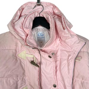 K&ouml;nnte beinhalten: Eine rosafarbene Steppjacke mit Kapuze und Rei&szlig;verschluss. Die Jacke hat einen wei&szlig;en Knebelverschluss und ein wei&szlig;es Etikett mit dem Text "sport futur courreges" auf der Vorderseite.