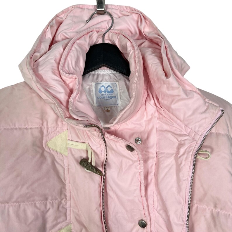 K&ouml;nnte beinhalten: Eine rosafarbene Steppjacke mit Kapuze und Rei&szlig;verschluss. Die Jacke hat einen wei&szlig;en Knebelverschluss und ein wei&szlig;es Etikett mit dem Text "sport futur courreges" auf der Vorderseite.