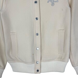 May include: Cream-colored varsity jacket with a button-up front, two front pockets, and a ribbed hem and cuffs. The jacket features a small, embellished design on the upper chest and silver-toned snap closures.