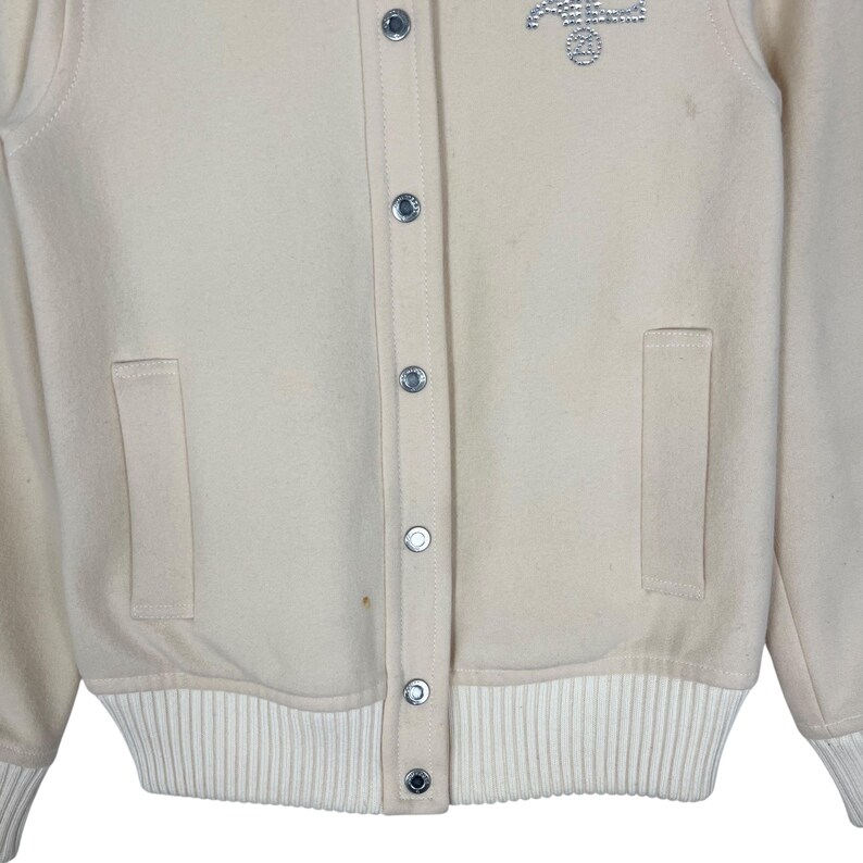 May include: Cream-colored varsity jacket with a button-up front, two front pockets, and a ribbed hem and cuffs. The jacket features a small, embellished design on the upper chest and silver-toned snap closures.